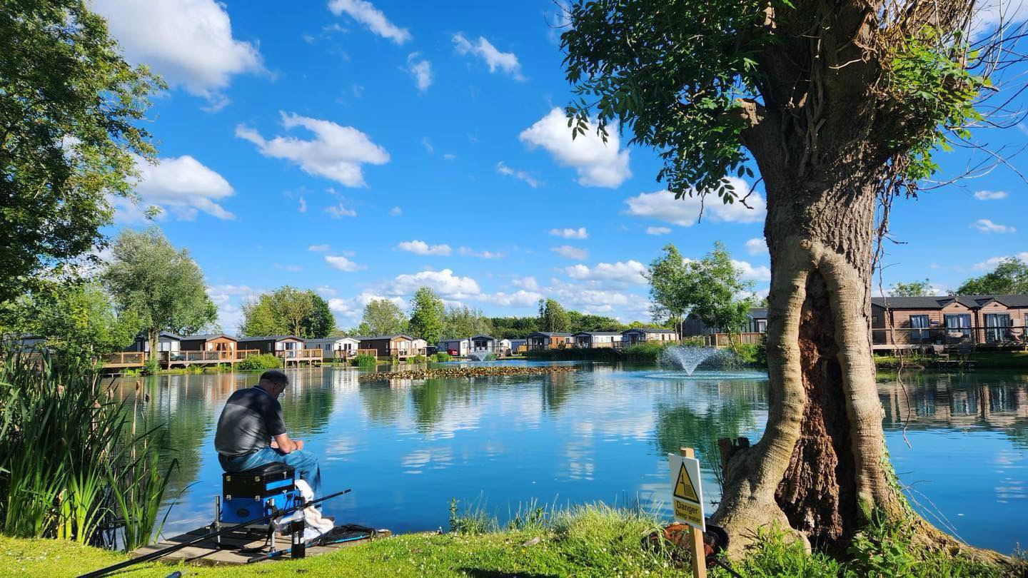 Man fishing beside the lake at Brickyard Lakes Country Park with luxury lodges overlooking the water, North Yorkshire holiday park.